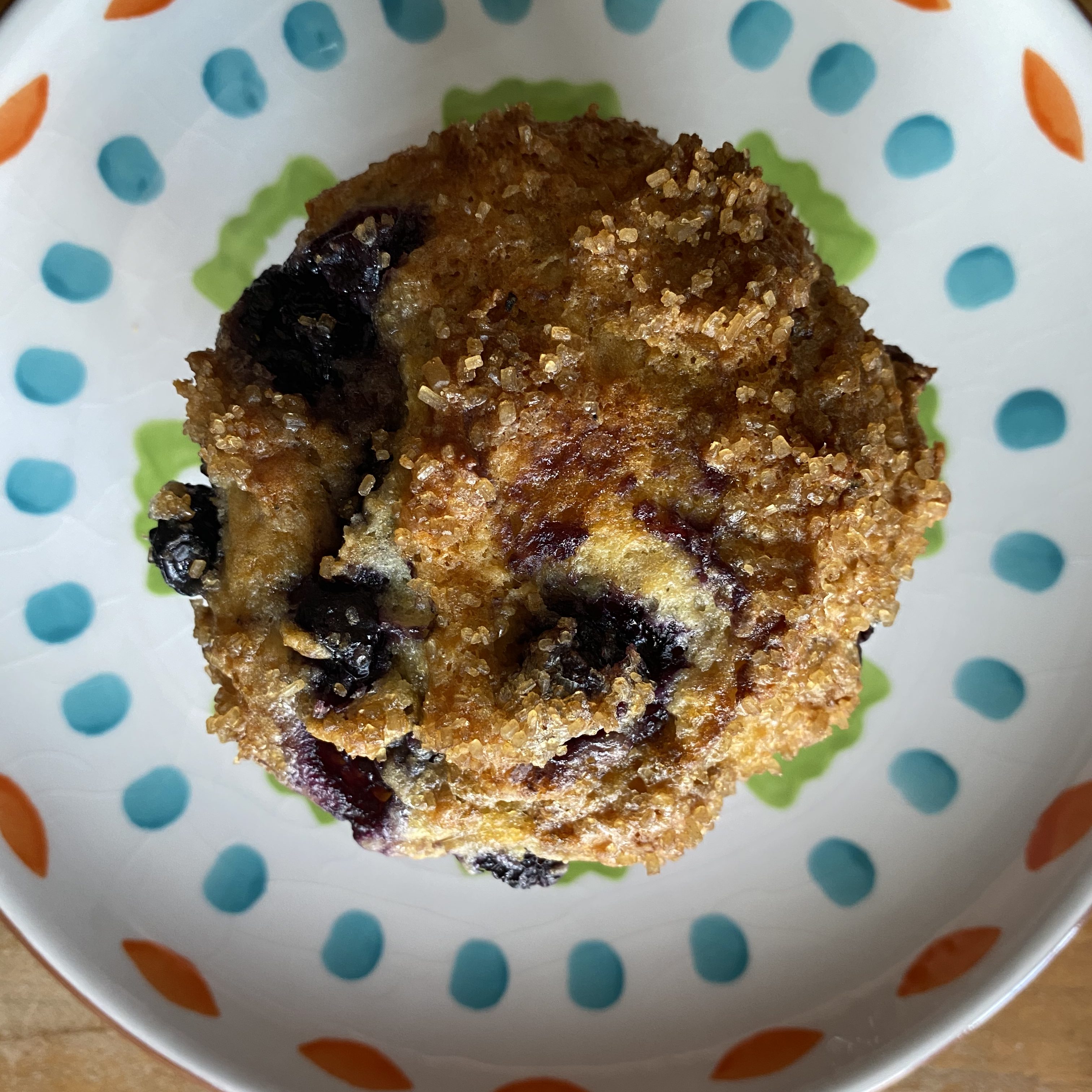 A top-down photo of a blueberry muffin on a colorful plate. The muffin is golden brown, with large sugar crystals on top and several dark blue and purple berries peeking out. The plate has a white background with greek, teal, and orange brushstrokes in an abstract pattern.