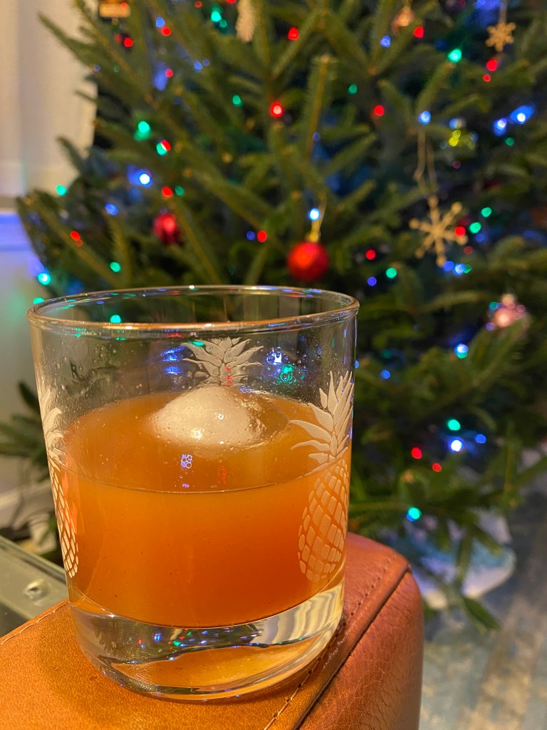 A tumbler with pineapple etching on it is resting on the arm of a brown leather couch. It contains a cloudy, apple-cider colored beverage and a round piece of ice. In the background is a blurry Christmas tree with lights and ornaments.