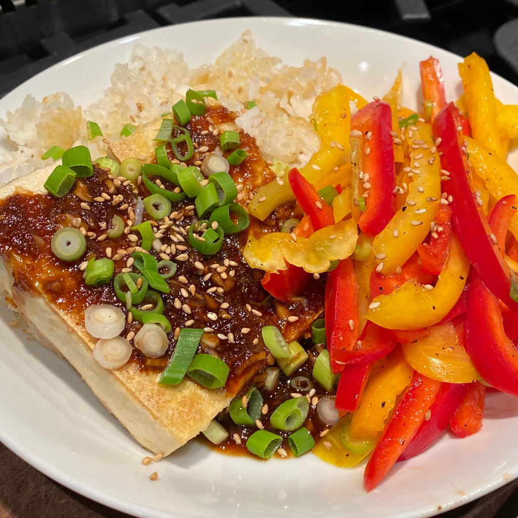 A rounded white plate almost entirely fills the frame of a square photo. On the right is a pile of sliced red and yellow bell pepper, at top is a pile of white rice with some lightly-browned cripsy bits, and at left are two slices of browned tofu covered in a dark brown sauce. Everything is scattered with toasted sesame seeds and sliced scallions.