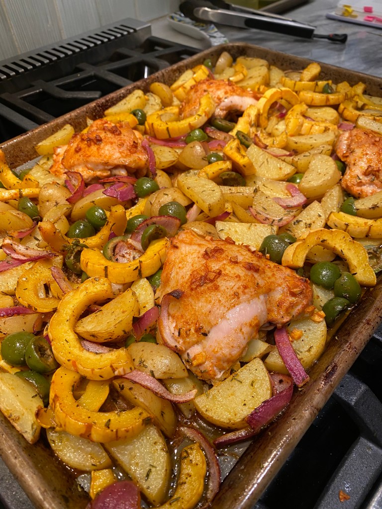 A sheet pan of cooked food is shown on top of a gas stove. The sheet pan contains roasted pieces of potato, delicata squash, red onion, green olives, and chicken thighs. The chicken thighs have an orange marinade nad there are flecks of spices on the veggies.