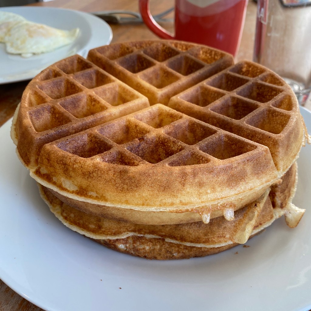 Two golden-brown waffles are stacked on a white dinner plate. In the background is another plate with two lightly-fried eggs and an orange and white pottery mug.