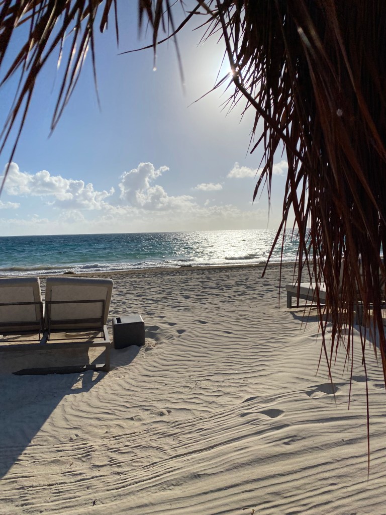 A view of the water taken from under a thatched umbrella. The Sky is a light blue, with the sun just hidden by some pieces of straw hanging down from the umbrella. The water is turquoise blue and set off by some fluffy clouds in the distance.