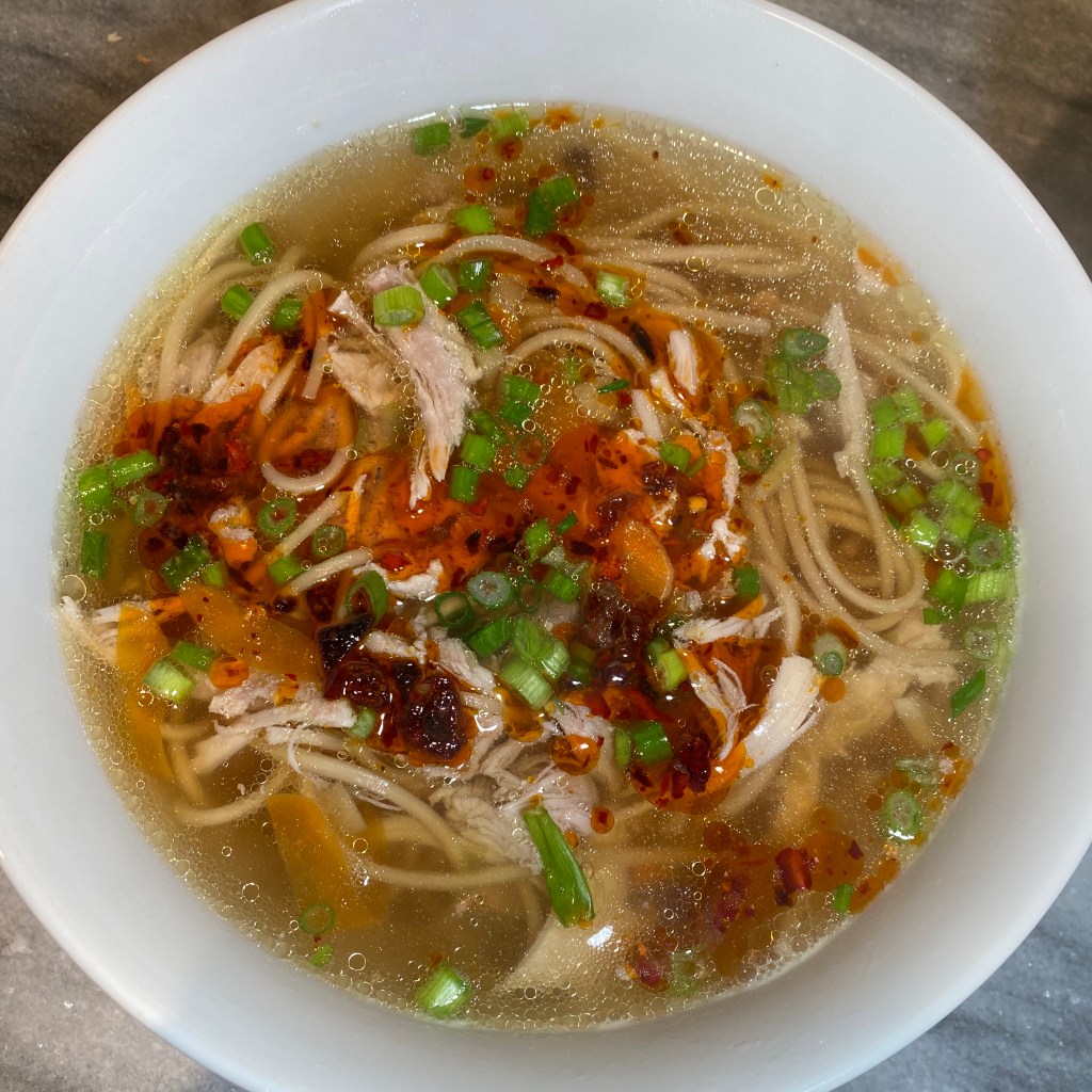 A white bowl almost fills the frame. It contains a soup with a light yellow broth, noodles, and pieces of chicken. It's topped with chopped scallions and a reddish condiment has been spooned over the top. Little bits of fat in the broth have caught the light, but in an extremely appetizing way.