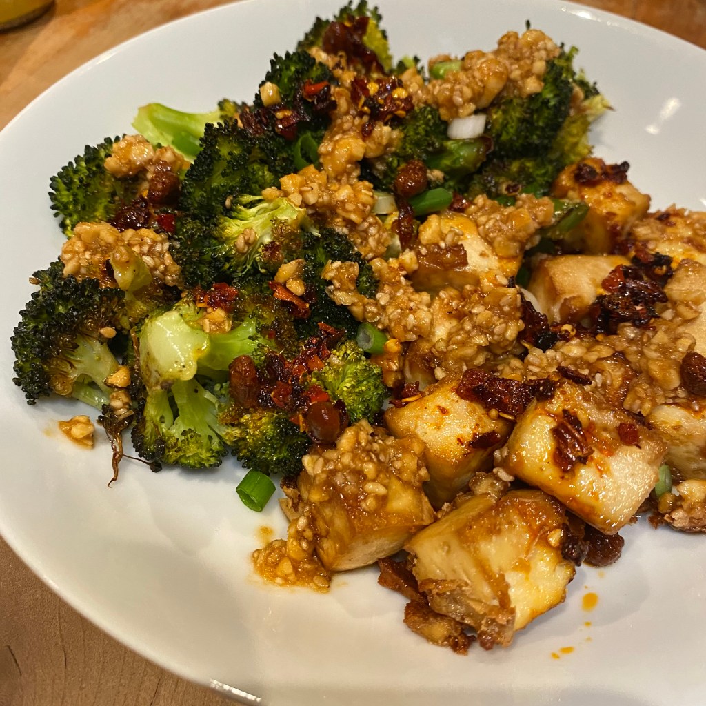 The frame is mostly filled by a shallow white bowl. Half of it is filled with roasted broccoli florets, and the other half with browned cubes of tofu. There are some chopped scallions scattered over, a healthy dose of sesame peanut pesto, and some chili crisp.