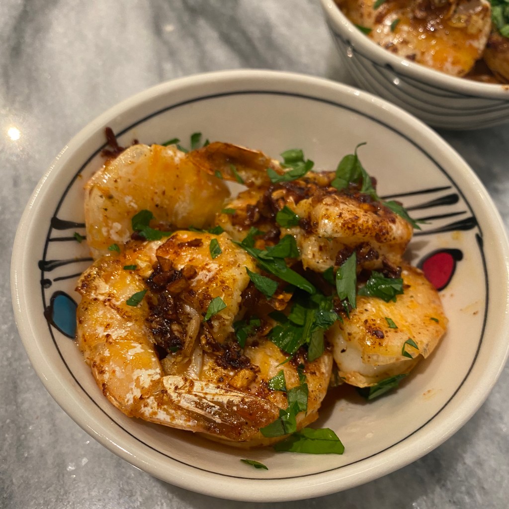 A small pile of cooked, shell-on shrimp rests in a cream-colored bowl with black, red, and blue accents. The shrimp are a peachy pink color, with some spots that are browned. There's a scattering of chopped parsley over the top, and you can see some pieces of garlic coated in a reddish sauce.