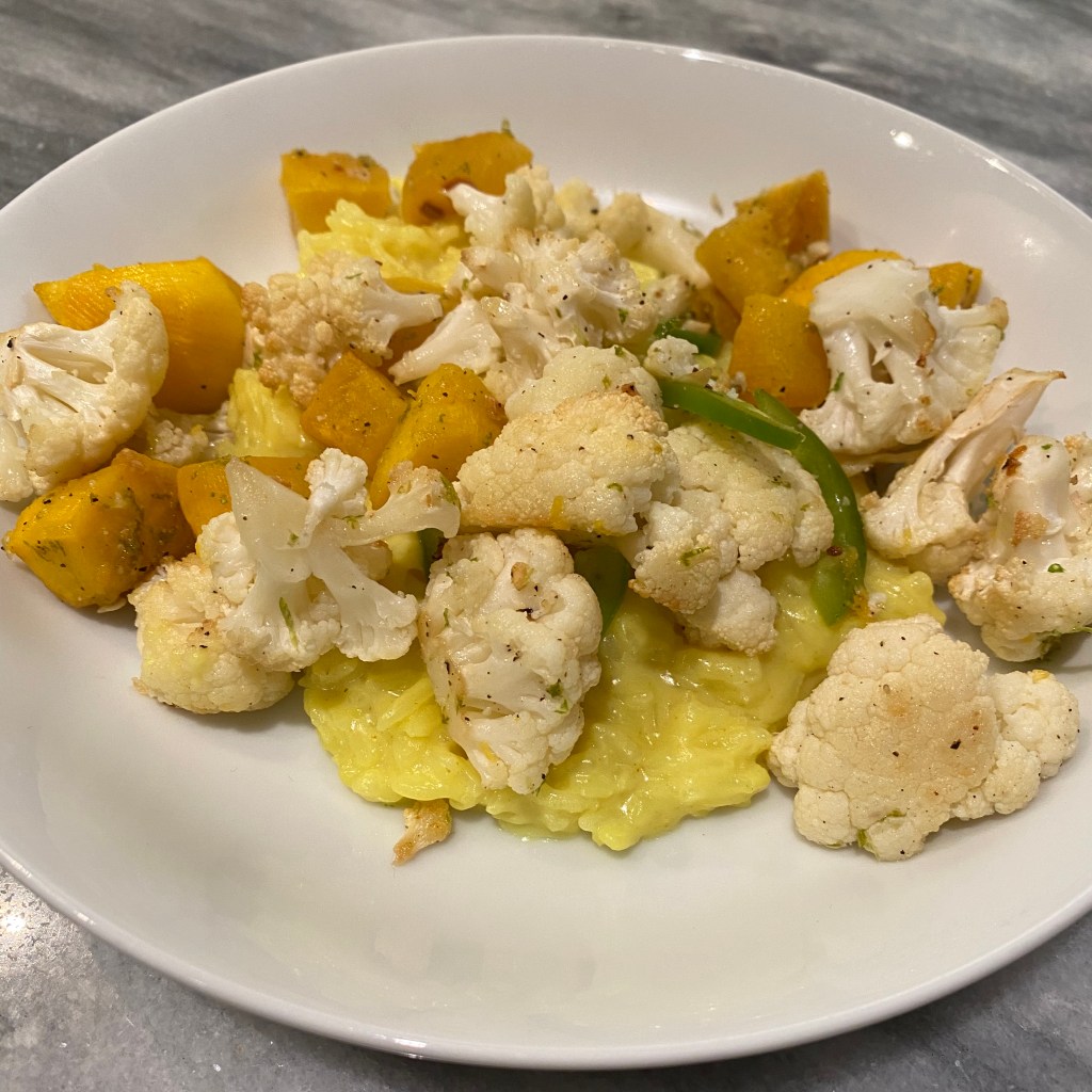 A shallow white bowl holds a big scoop of creamy yellow rice. It's topped with roasted cauliflower florets and pieces of squash. There are a couple of slices of jalpeno peeking out.