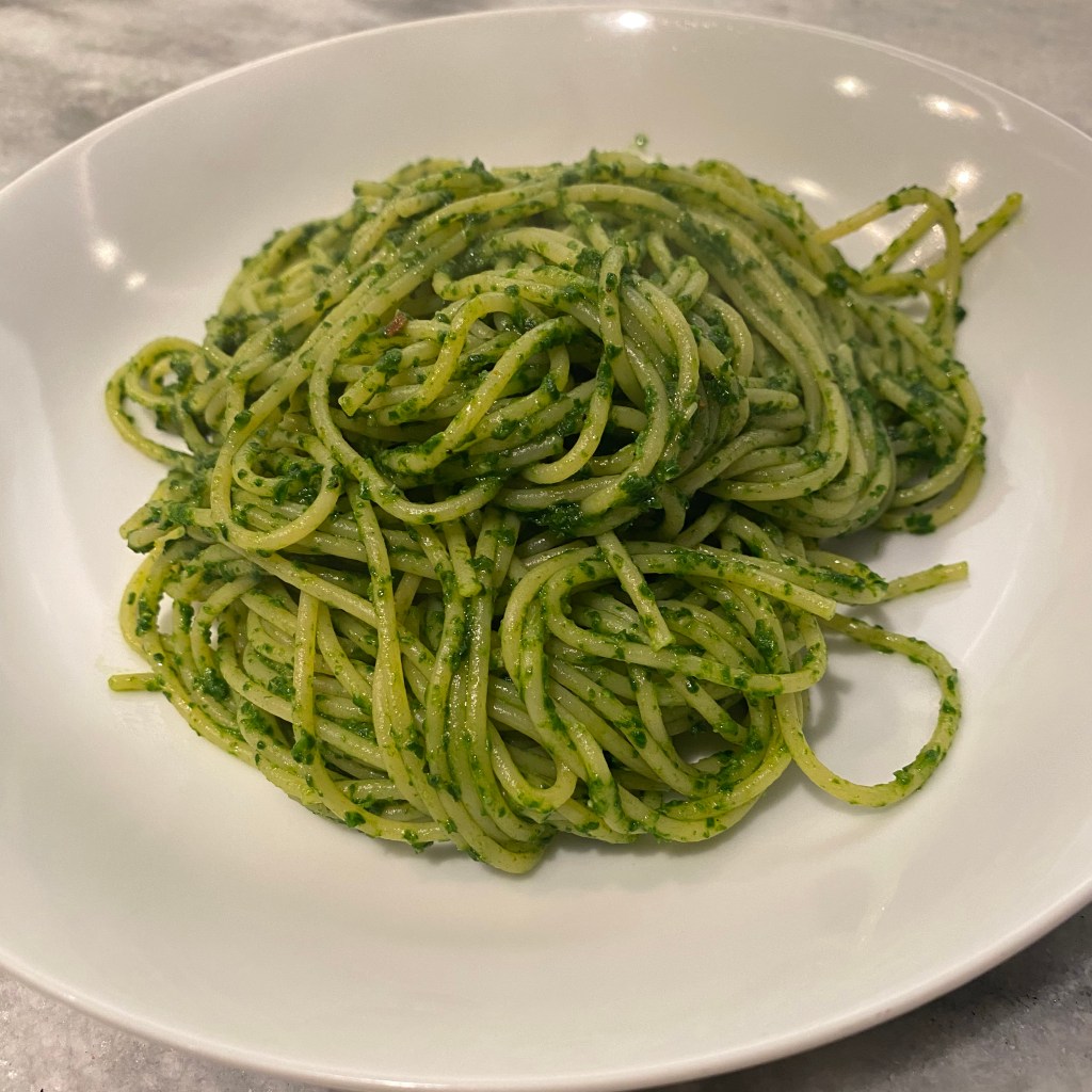 A shallow white bowl nearly fills the frame. A tangle of thin spaghetti is heaped in the middle. The pasta is coated in a green sauce with lots of flecks of spinach throughout.