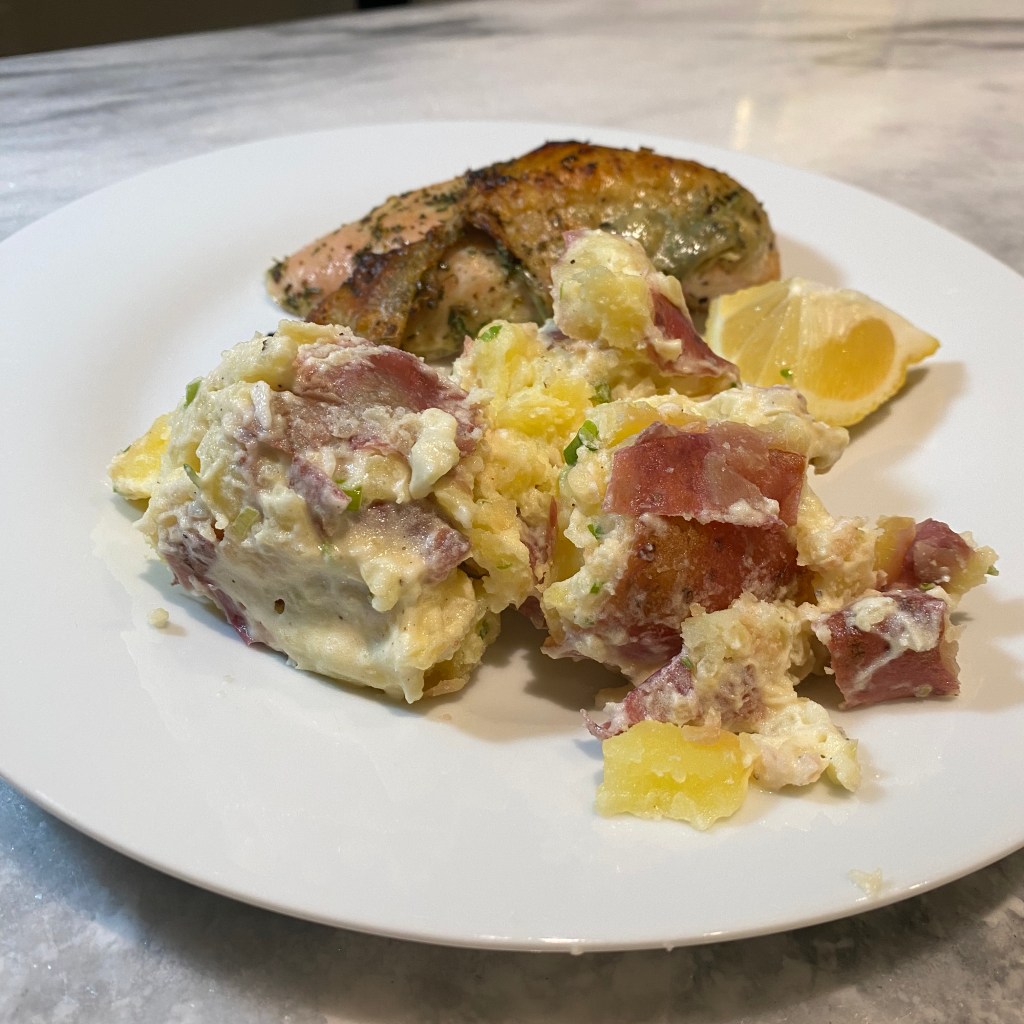 A pile of cooked and partially mashed potatoes sits at the front of a white plate. The potatoes have a white sauce with flecks of scallion. A small wedge of lemon and a roasted skin-on chicken breast covered in herbs are on the back of the plate.