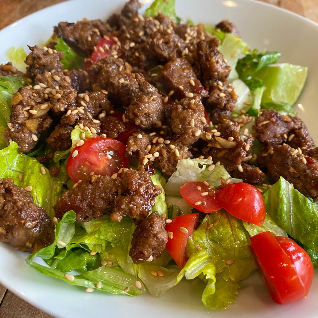 A pilot of chopped romaine lettuce rests in a shallow white bowl. The lettuce is very lightly dressed, just enough to catch the light, and topped with chopped cherry tomatoes, toasty pieces of browned ground beef, and a shower of toasted sesame seeds. A wedge of lime peeks out from the back right.