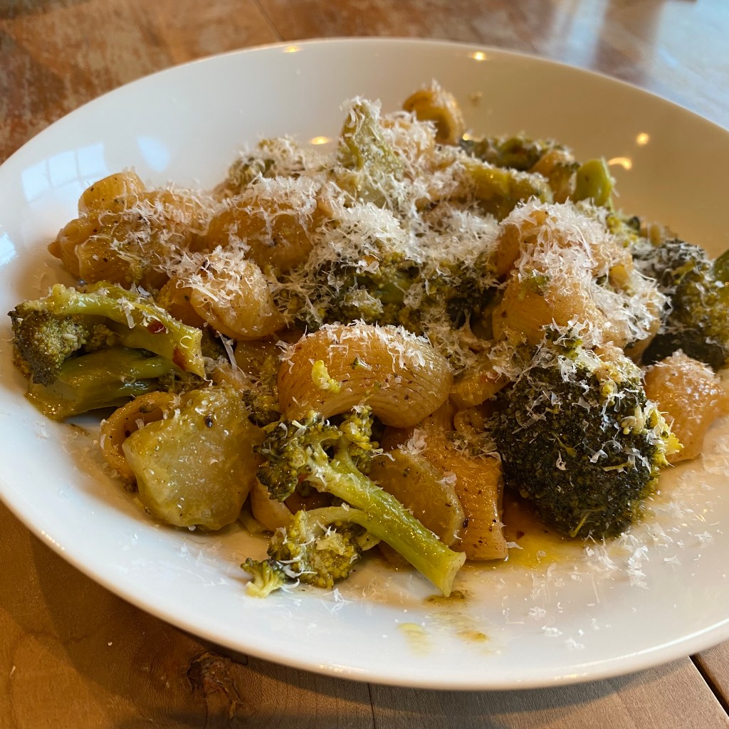 A shallow round plate contains a pile of broccoli and pasta that is mostly obscured by parmesan. The broccoli is an olive green and everything has a somewhat golden brown cast, which is not due solely to the poor lighting.
