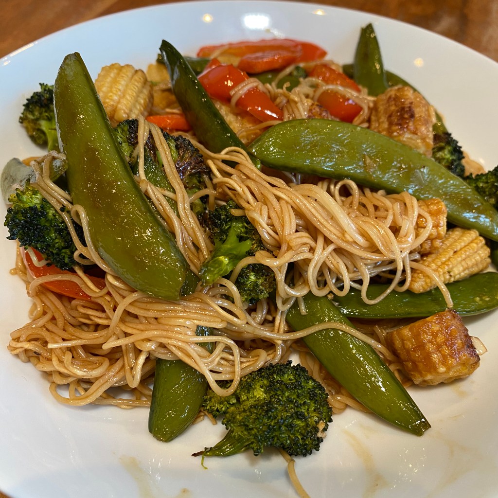 A shallow white bowl contains a pile of thin noodles covered in a light brown sauce. There are sugar snap peas, broccoli, baby corn, and red bell peppers tangled in the noodles. All the veggie shave signs of being lightly roasted. 