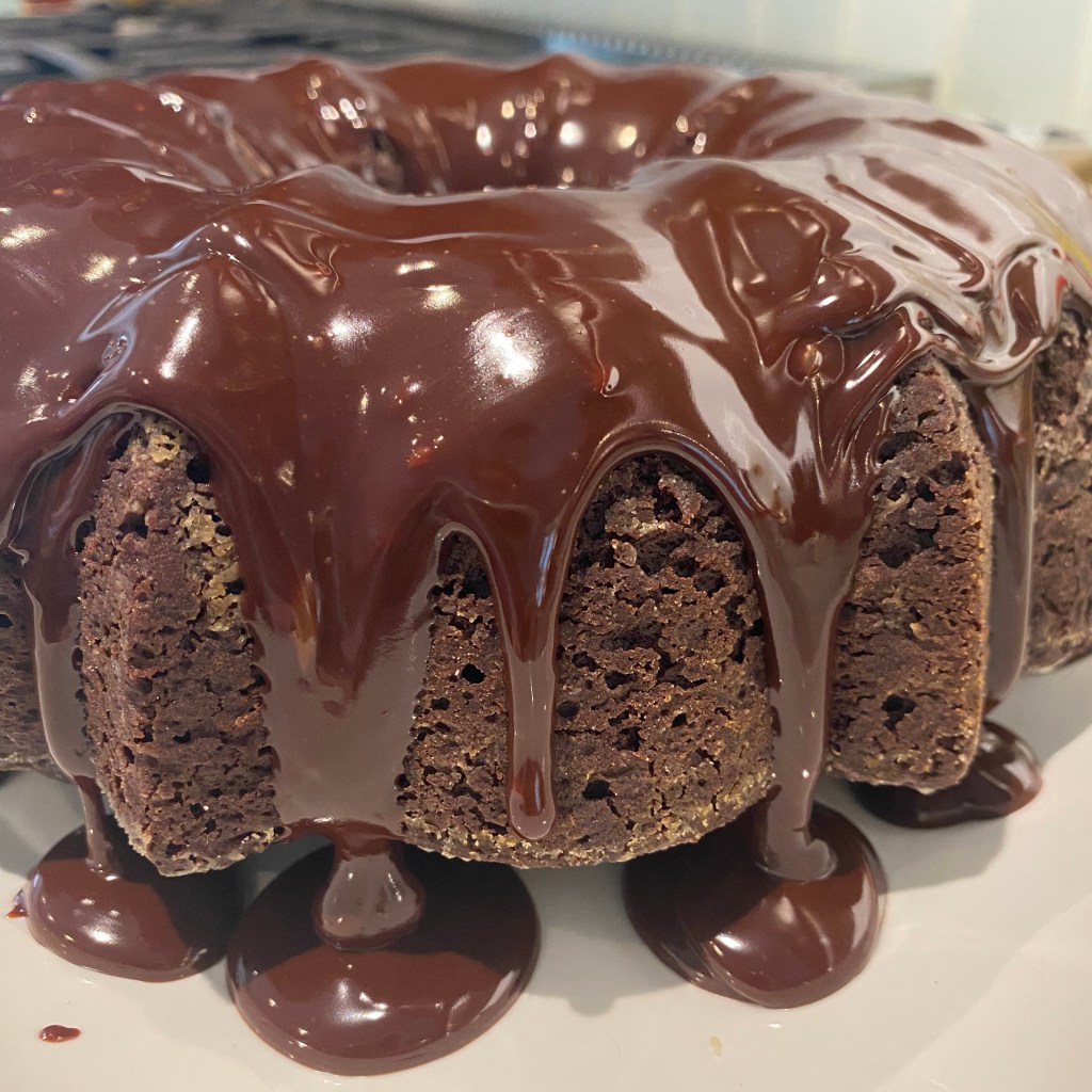 A side view of a chocolate bundt cake with chocolate ganache dripping off of it. The cake appears to be hovering over a white platter, with chocolate pooling underneath it. The ganache is shiny, which distracts you from a few spots where you can see the flour used to coat the pan.