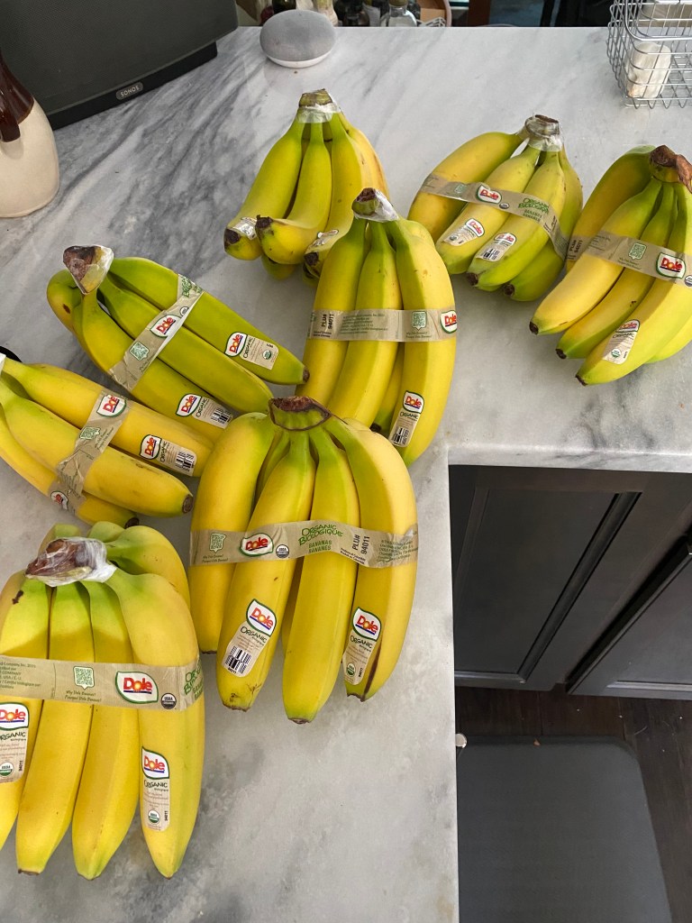 Eight bunches of bananas are shown strewn along the inside corner of an L-shaped countertop. They are mostly very close to ripe, and all have brown, Dole-branded tape holding them together.