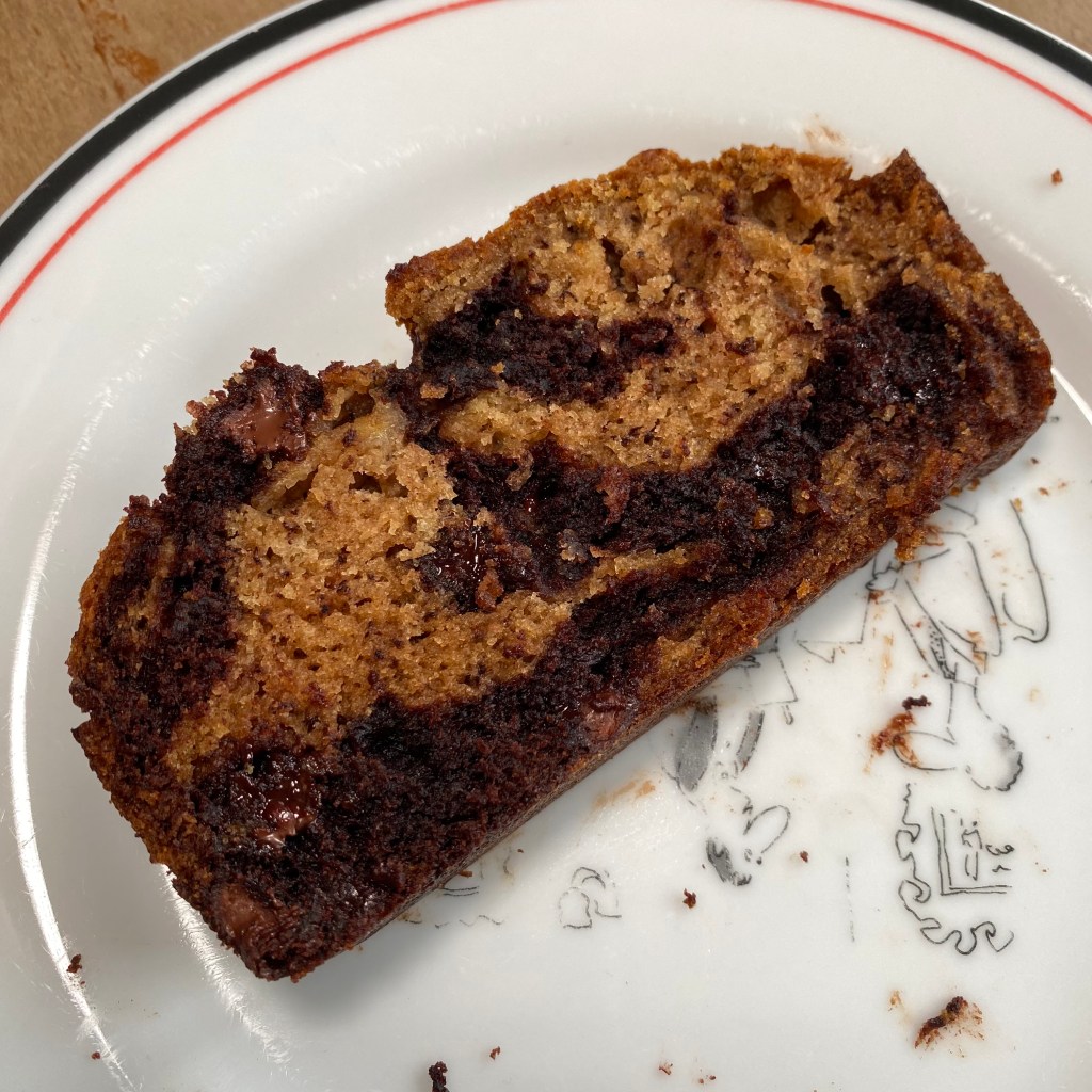 A slice of banana bread marbled with chocolate sections rests on a white plate with part of a New Yorker cartoon showing. The chocolate sections are almost black, and in several spots you can see the shine of melted chocolate chips that haven't quite solidified yet.