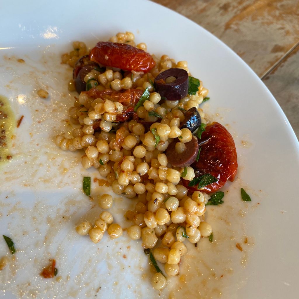 A white plate with the last few forkfuls of a serving of pearl couscous mixed with black olives and roasted halves of grape tomatoes. There are flecks of parsley throughout, and the left side of the plate has streaks of sauce. 