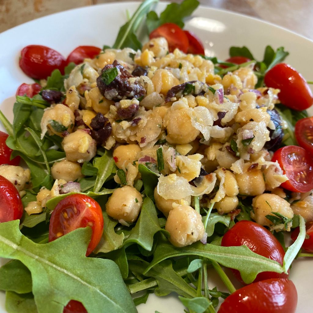 A shallow white bowl holds a pile of baby arugula. It's covered with a scoop of chickpeas, with black olives, flecks of parsley, and red onion scattered throughout. Several halved grape tomatoes are surrounding it.