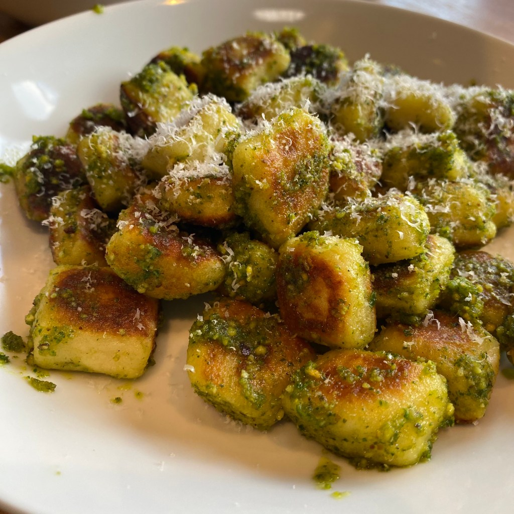 A pile of gnocchi rests in a shallow white bowl. They're pillow-shaped, and the larger sides are golden brown. It's all coated in a slightly chunky green pesto, and showered in finely grated parmesan.