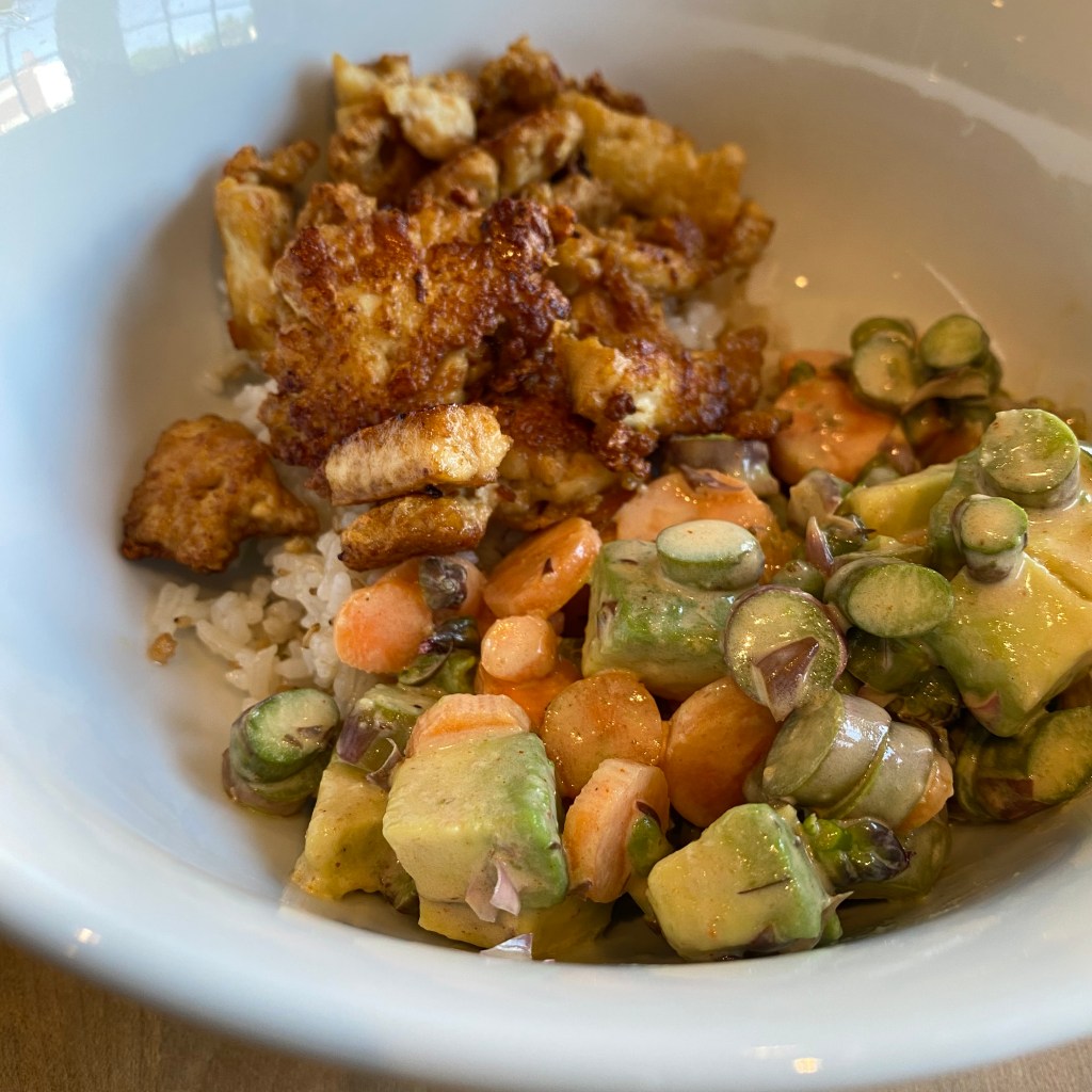 A white bowl holds a pile of white rice, which is almost entirely obscured by a pile of fried, irregularly shaped tofu at the back. At front is a salad of carrot and asparagus coins with cubes of avocado, coated in a light, beige-colored dressing.