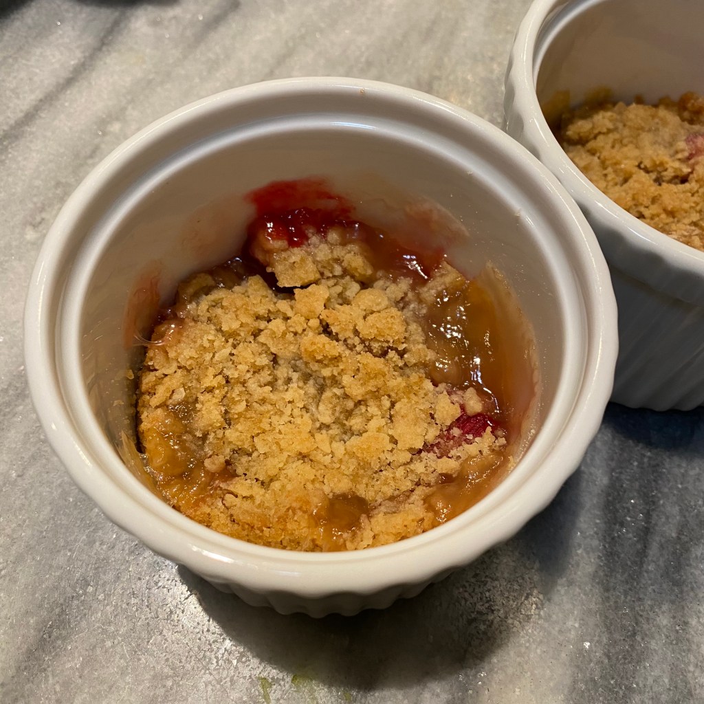 A round white ramein is shown from above. The top is mostly covered in crumbly, golden brown topping. Around the edges you can see some green and pink pieces of rhubarb that have baked themselves into jam. Another ramekin is visible in the top right.