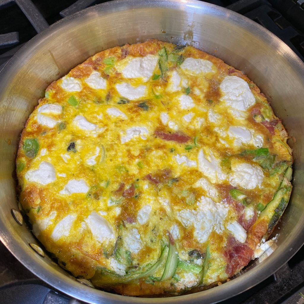 A round, stainless steel skillet fills the frame. It holds a yellow fritatta with golden brown edges, and some light browning in the middle. There are many white goat cheese islands, with a few peeds of green asparagus and pink prosciutto.