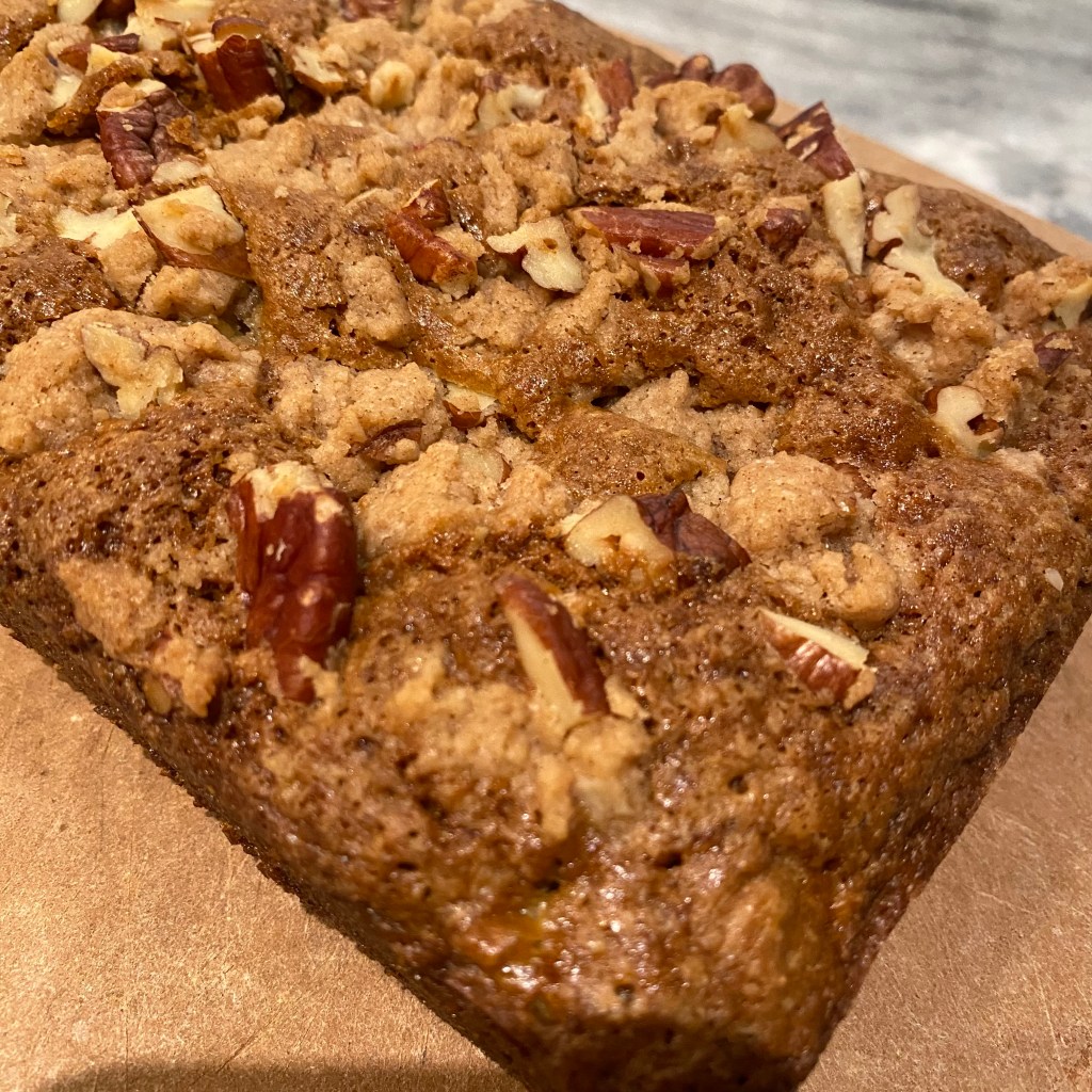 A top view of a squat brown loaf, with toasted pecans and areas of lighter-brown streusel. The loaf has been placed on a brown cutting board, and some whitish grey countertops are visible behind it.
