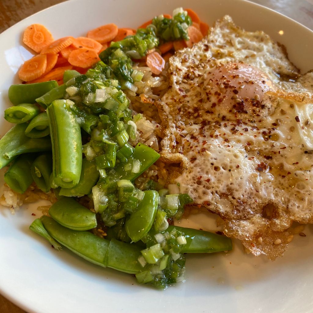 A shallow white bowl fills the frame. At right there's a fried egg, with browned lacy edges. It's covered in red flakes of gochugaru and some toasted sesame seeds. At front left are some roughly chopped snap peas, with carrot coins behind. Some brown rice is peeking out between the eggs and veggies, and there's a trail of diced scallion coated in oil down the middle of the plate.