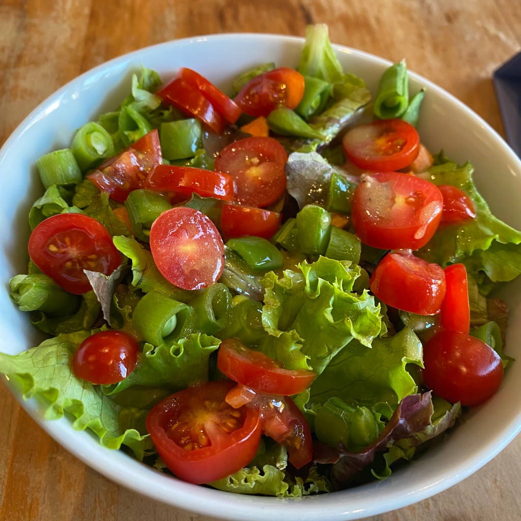 A white bowl holds a fresh green salad. There is pieces of curly green lettuce, chopped grape tomatoes, small coins of carrot, and chopped snap peas. It's been drizzled with salad dressing so there are bigs of oil catching the light.