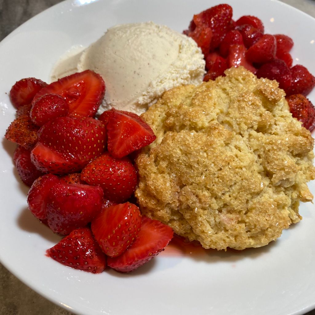 A shallow white bowl contains, front right, a craggly, golden-brown shortcake studded with large sugar crystals. At the back left is a scoop of vanilla ice cream flecked with vanilla bean bits. The top right and bottom left are filled with halved strawberries, and you can see a little puddle of strawberry juice gathering underneath it all.