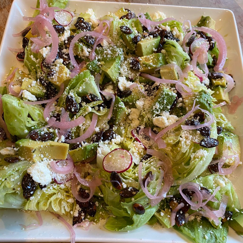 A square white platter holds small wedges of iceberg lettuce, liberally covered in crumbled white cotija cheese, rounds of red radish slices, diced avocado, baby pink pickled onions, and dark brown toasted pumpkin seeds.