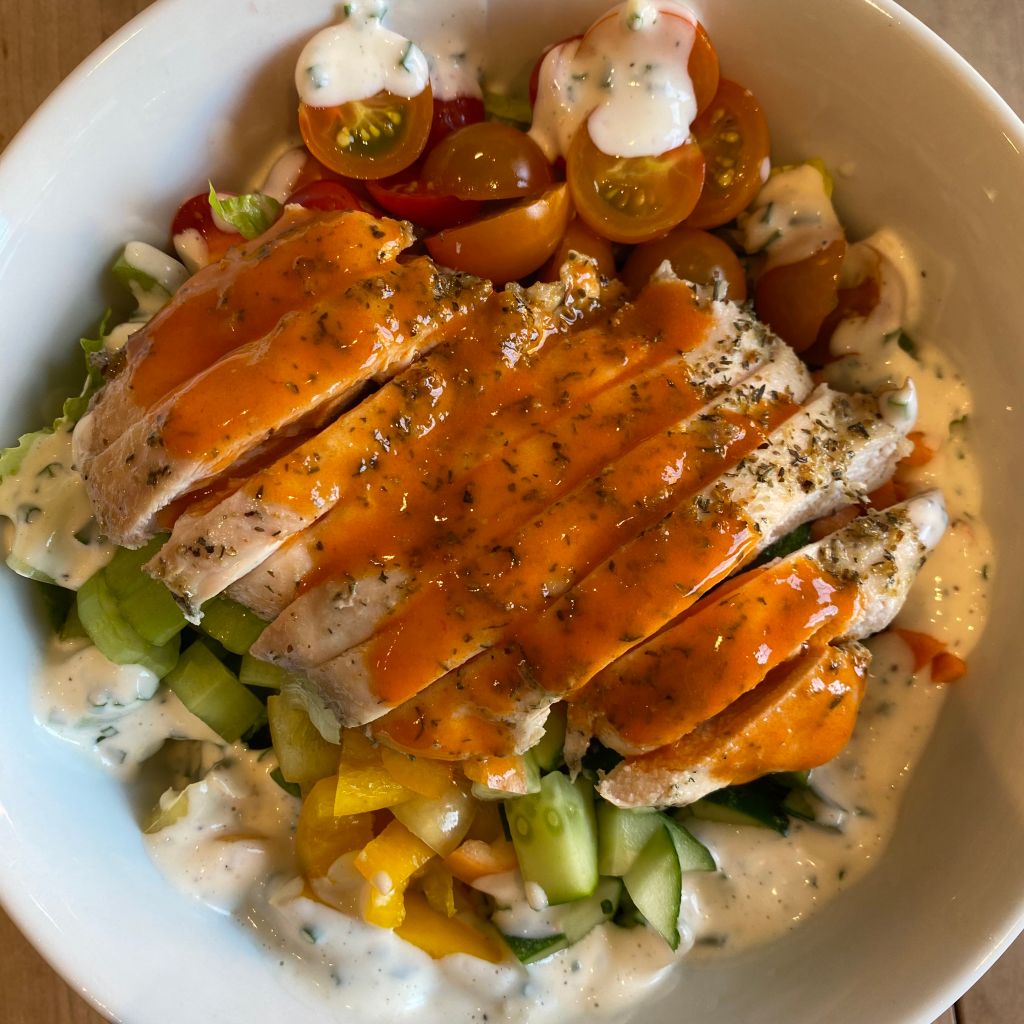 A sliced chicken thigh, flecked with spices and doused in an orange sauce, rests on top of a bed of romaine lettuce you can barely see. Around the edges of the chicken are chopped yellow peppers, celery, orange and red cherry tomatoes, carrots and cucumbers. They're all drizzled with a white dressing with a generous amount of chives. 