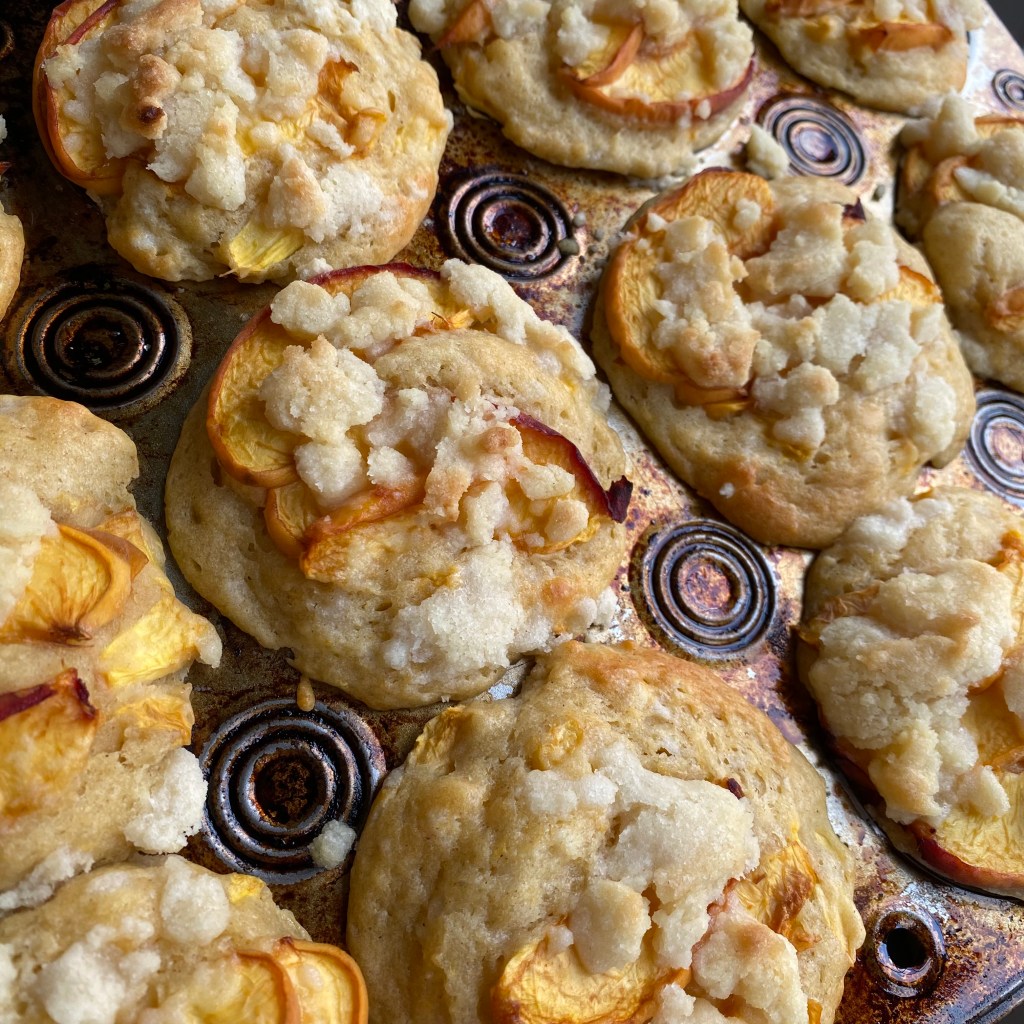 A muffin tin with a lot of brown and yellow patina on it is full of lightly golden brown muffins. Each of them is topped with slightly wrinkled pieces of yellow peach, with pieces of the orange skin, and some beige crumble topping.