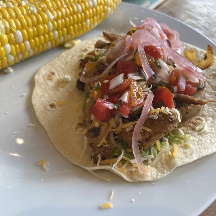 A white plate holds a yellow ear of corn behind a flour tortilla layered with food. Peeking out from teh bottom is a smear of guacamole. You can see some chunks of chicken and pieces of browned onion with pieces of shredded cheese poking out. There's some diced pieces of red and orange tomatoes coated with cilantro and mixed with pieces of white onion, and then some ribbons of light pink pickled red onions. It is definitely too much food for the size of the tortilla but we made it work.