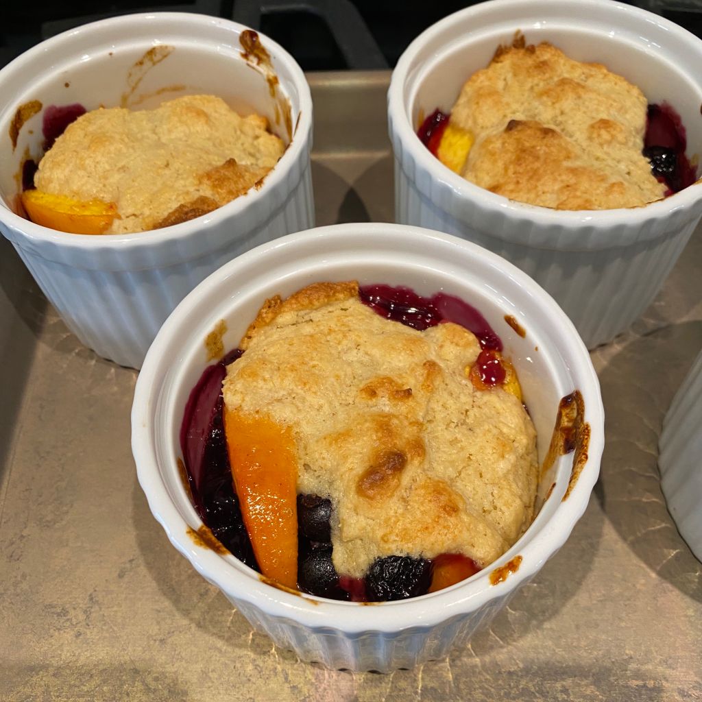 Three round white ramekins sit on a silver baking tray. The one in the front  has an orange slice of peach peeking out from under a blob of golden brown cake. There are several dark colored blueberries, next to the peach slice, and all around the edges you can see a purple jammy liquid.