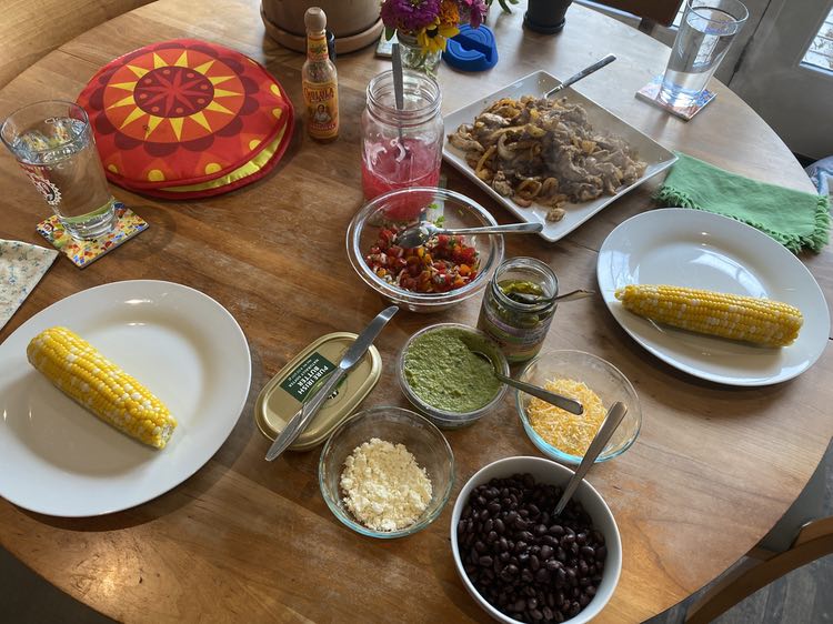 A round wooden table is cluttered with dishes. They include: two white dinner places, each with an ear of corn; a red and yellow tortilla warmer; a quart jar of pink pickled onions; a glass bowl with red pico de gallo; a tub of guacamole; a jar of pickled jalapenos; two small glass bowls, one with yellow and white shredded cheese, the other with white crumbled cotija; and a white bowl of black beans. Most of the bowls and jars have a spoon or fork stuck in them. There are also mismatched napkins (one green, one beige with a tiny floral print); and colorful mosaic pattern coasters with pint glasses of water. In the background there's a bottle of Cholula hot sauce, a small floral arrangement, and some planters. 