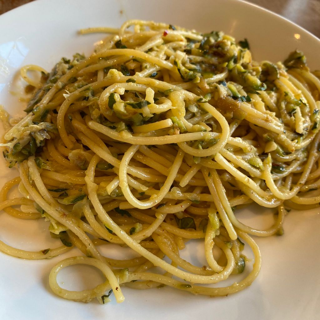A tangle of spaghetti sits in a shallow white bowl. Flecks of green zucchini are mixed throughout, 