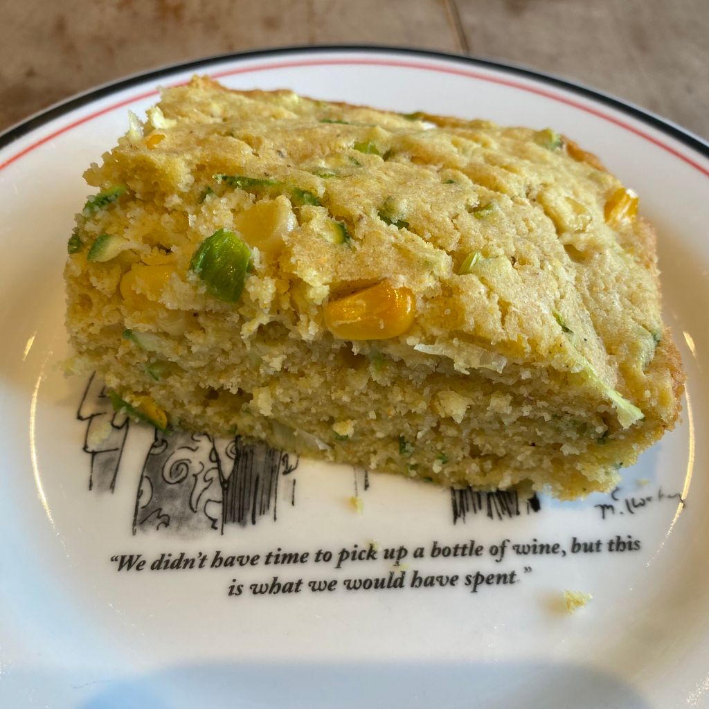 A rectangular piece of yellow cornbread sits on a white plate with a red and black striped rim. The cornbread is speckled with green bits of zucchini and chives, and whole pieces of yellow corn. It's covering up a cartoon, but you can still read the caption: "We didn't have time to pick up a bottle of wine, but this is what we would have spent."