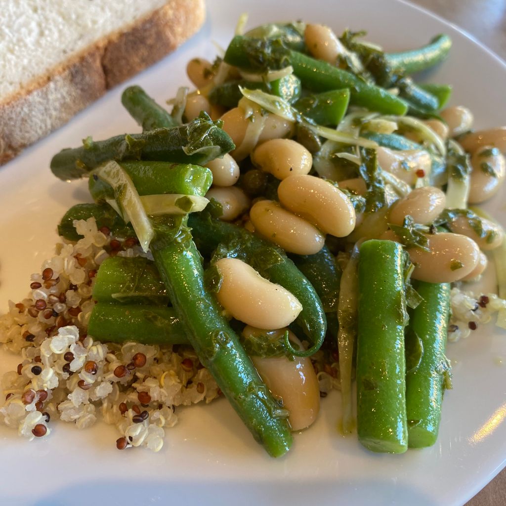 A pile of bright green beans and beige cannellini beans rests on top of some red and white quinoa. The beans are coated in flecks of dark green basil and slices of fennel.