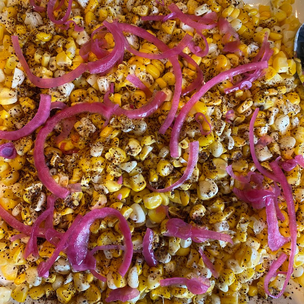 Yellow and white pieces of corn fill the frame. They're topped with dark brown flakes of chile powder and bright pink pieces of pickled red onion. 