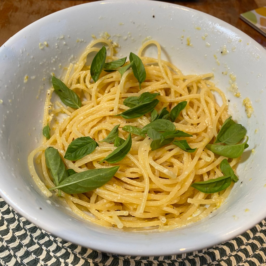 A large white bowl holds a tangle of spaghetti with a handful of small basil leaves scattered over the top. The pasta, and the sides of the bowl, have flecks and small blobs of parmesan.