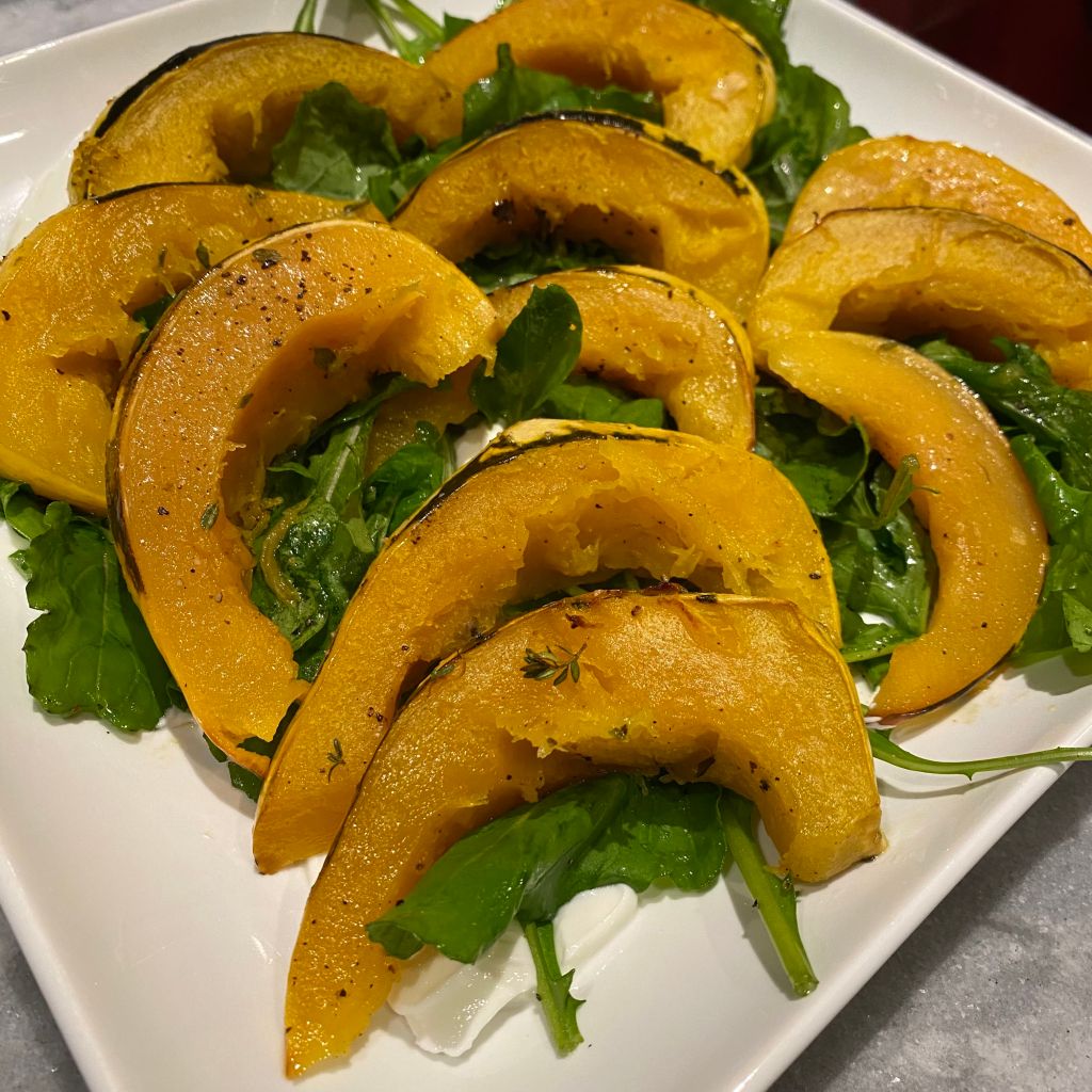 A square white serving platter holds golden wedges of roasted acorn squash that are neatly lined up on top of a bed of green arugula. There are swooshes of white yogurt peeking out from under the greens.
