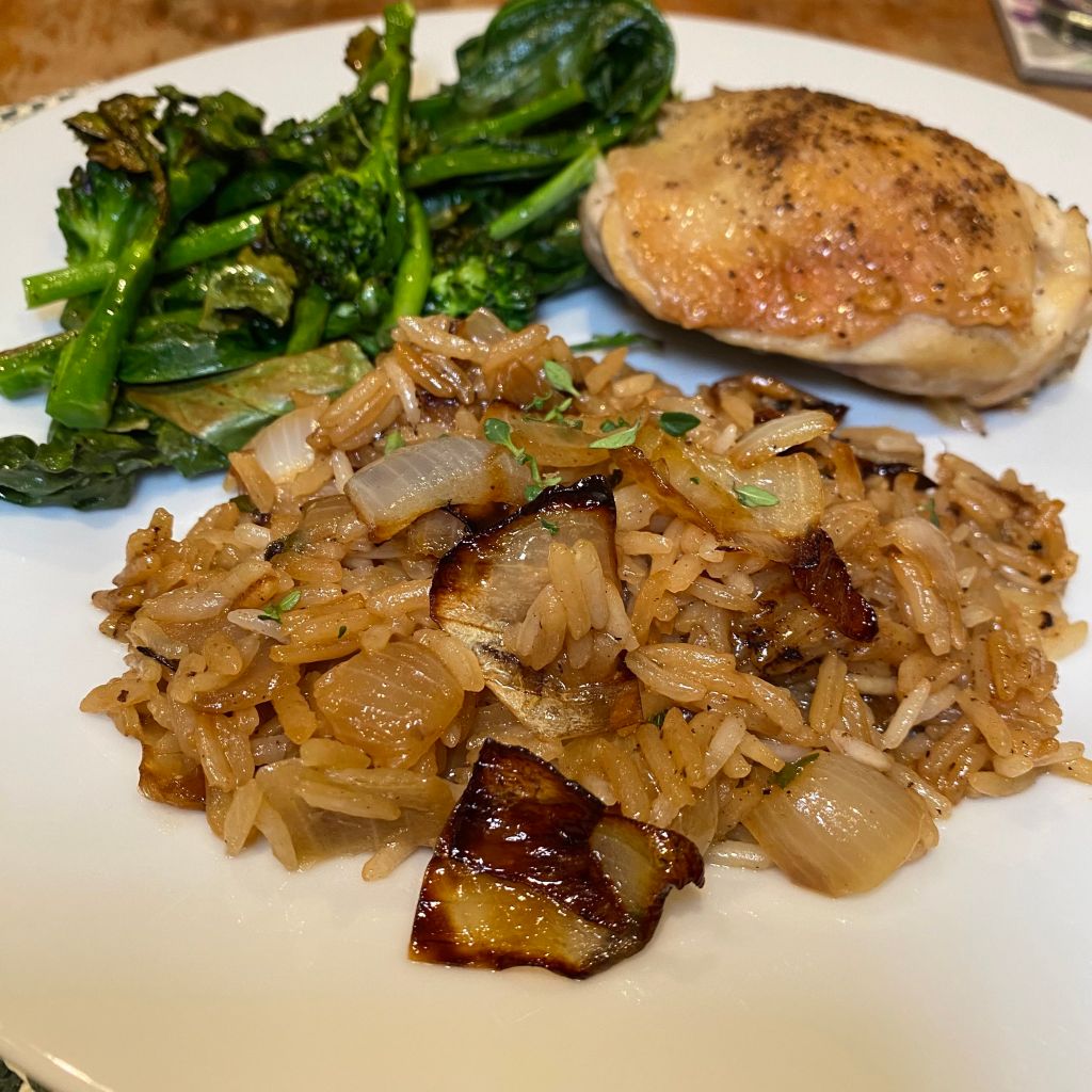 A scoop of golden brown rice studded with chunks of cooked onion sites at the front of a white plate. Behind it is a pile of dark green sprouting broccoli and a golden chicken thigh.