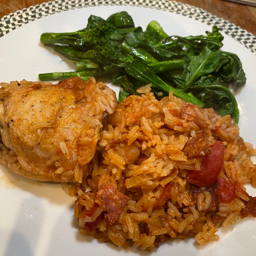 A white plate holds a pile or reddish rice, with pieces of browned chorizo and red tomatoes scattered throughout. To the left of the rice is a golden brown chicken thigh. Behind them both is a pile of long spears of deep green sprouting broccoli.