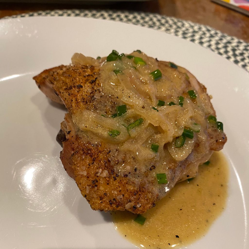 A golden brown chicken thigh rests on a white plate. It's smothered with a brown sauce of sliced shallots with flecks of green chives. Some crispy bits of skin are peeking out from under the sauce, and there's a small puddle forming underneath.