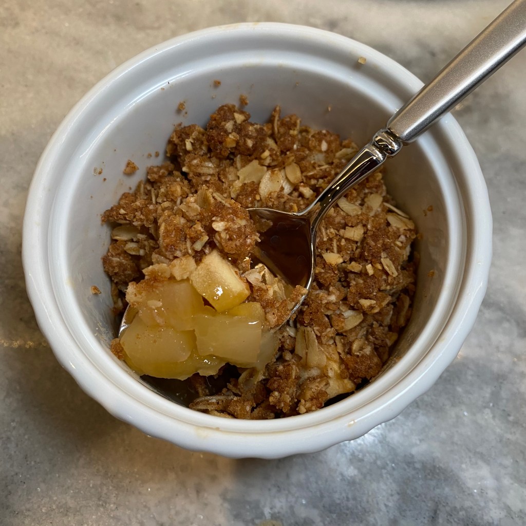 This is a top-down shot of a round white ramekin rests on a grey marble counter. A spoon rests in the ramekin, holding a bite of apple crisp. The apples are golden and the crisp is a dark brown, flecked with oats.