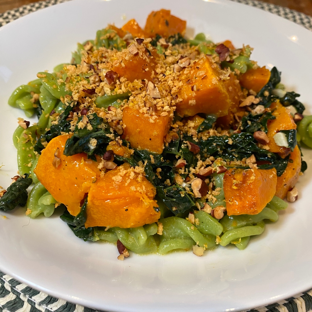 A shallow white bowl holds a pile of pasta. Peeking out from the bottom are pieces of light green rotini. Piled on top are orange chunks of squash and dark green pieces of kale. The whole thing is showered with golden brown panko and chopped toasted pecans.