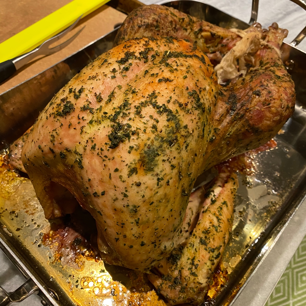A golden-brown turkey covered in green herbs rests in a silver roasting pan with brown drippings below it. it looks delicious because it is.