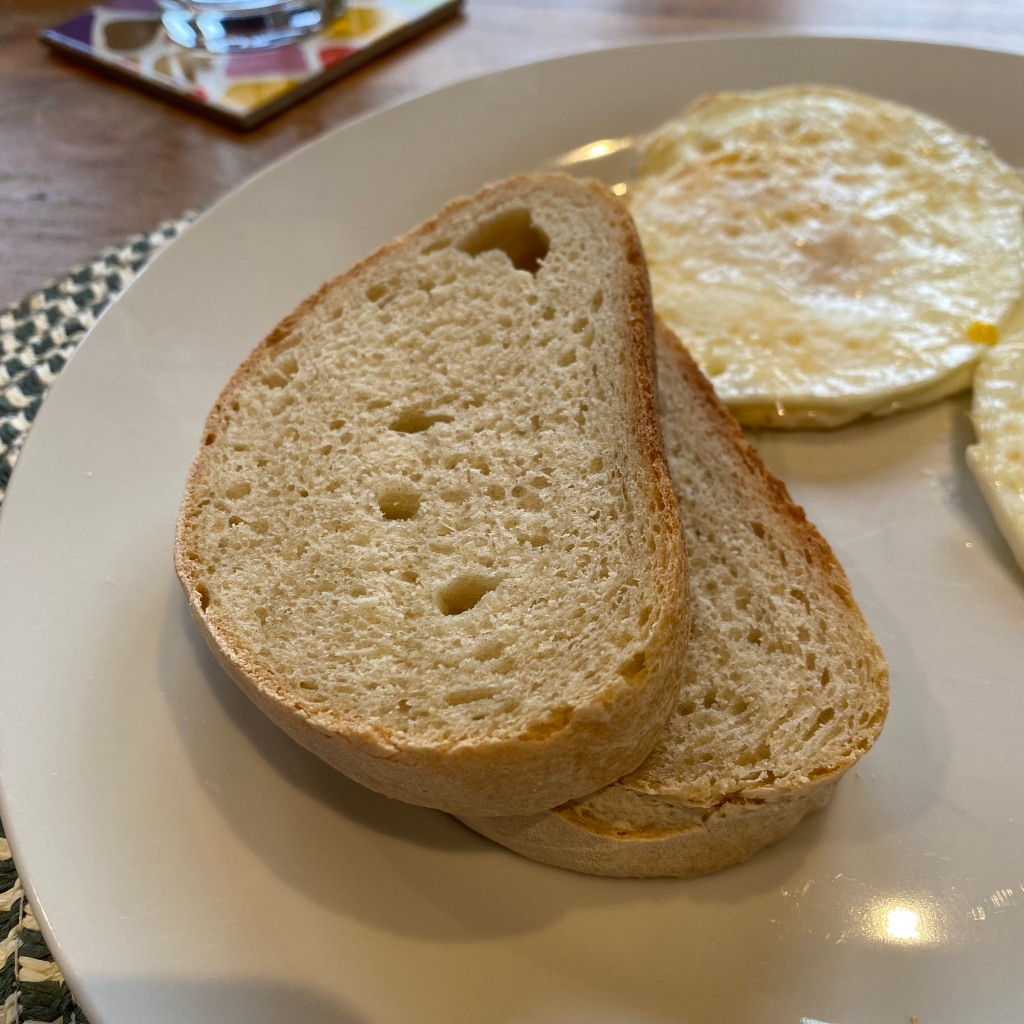 Two slices of white bread a stacked on a white plate with a fried egg in the background. The bread has a very light brown crust, and a mostly even scattering of small holes, with four larger ones going through the middle. 