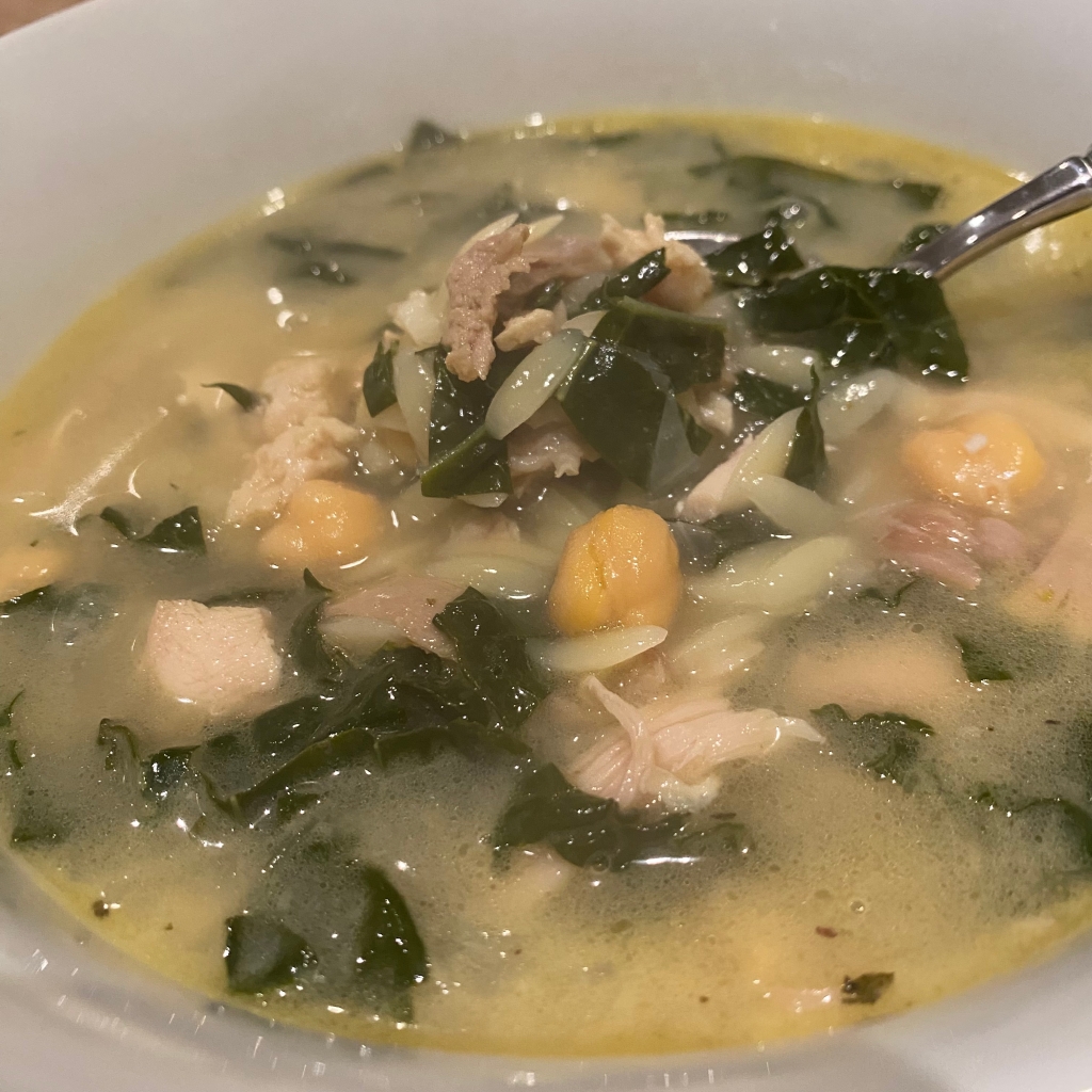 A white bowl holds a serving of soup. It has a cloudy yellow broth, and there are chickpeas, small pieces of dark and white meat turkey, white pieces of orzo, and dark green pieces of kale. The handle of a spoon gleams from the upper right corner.