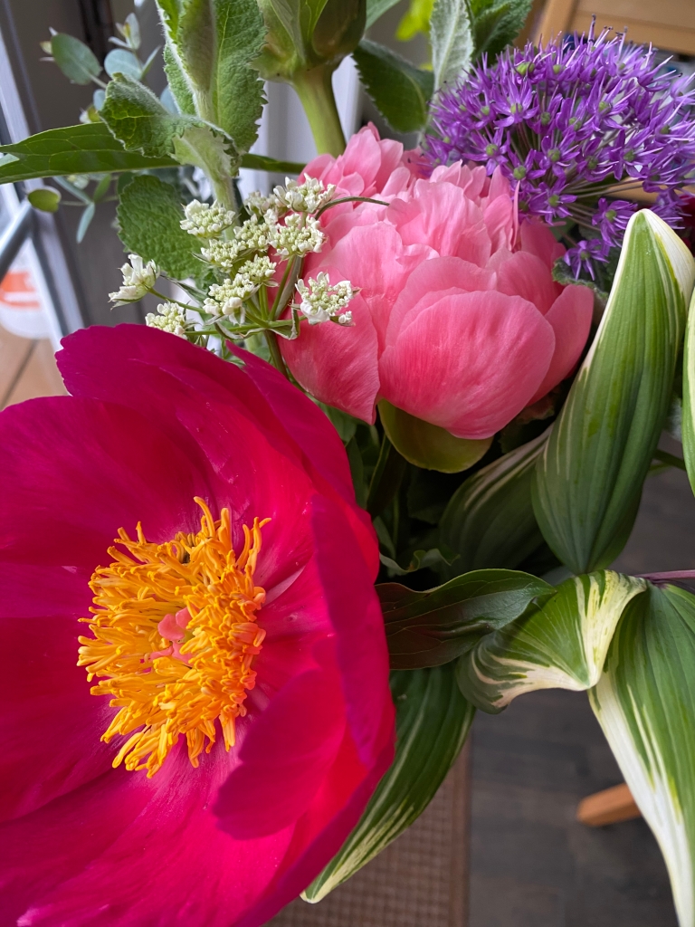 A bouquet featuring a dark pink peony in full bloom, and a lighter pink, fluffy peony behind. There are also some greens and filler flowers in white and purple.