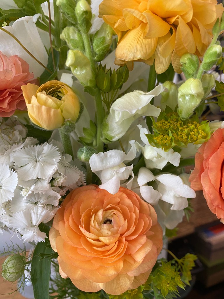 A close up of a bouquet showing white snapdragons, peach and yellow ranunculus, yellow yarrow, and some other white and green flowers peeking out on the sides.